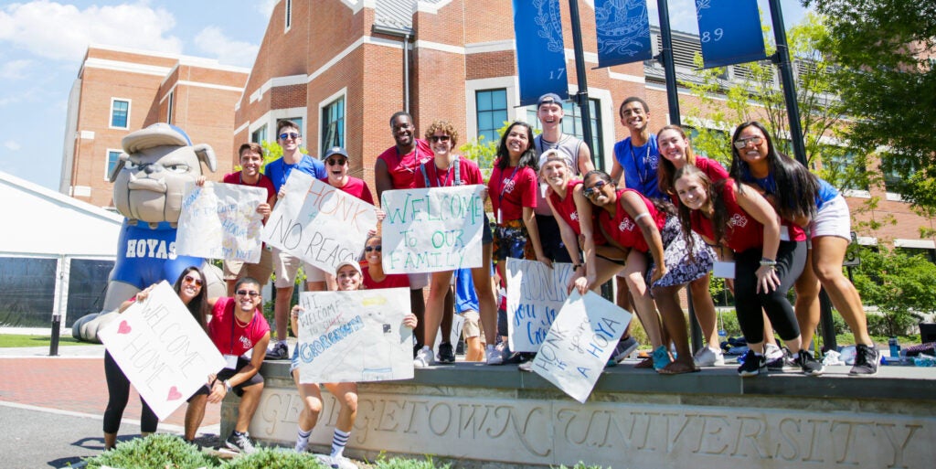 Students in red T-shirts hold signs outside welcoming new students to Georgetown