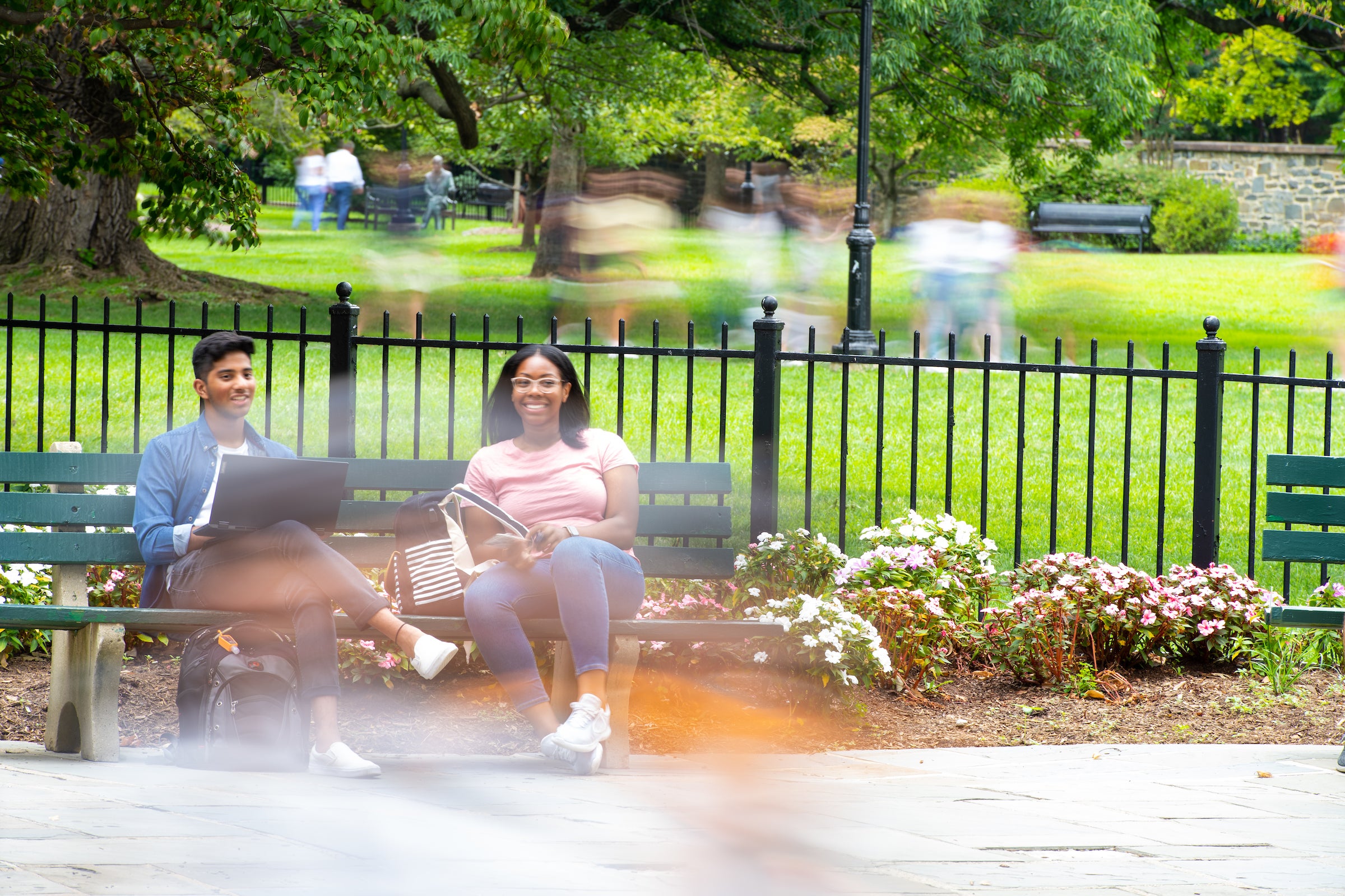 Two students sit on a bench while students walk out of focus around them